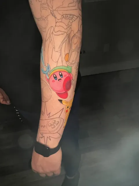 Anime forearm tattoo of Kirby wearing a green cap, placed among the outlines of a video game sleeve in progress