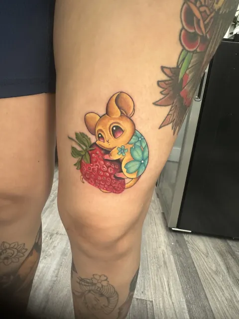 Anime thigh tattoo of a small yellow mouse holding a strawberry beside a blue flower
