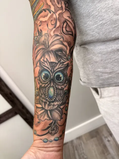 Healed color forearm tattoo of an owl with jewel-blue eyes among lilies and scrollwork