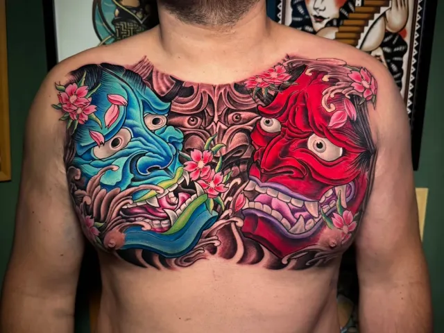 Full-chest neo-oriental tattoo of two hannya masks, one blue and one red, surrounded by cherry blossoms