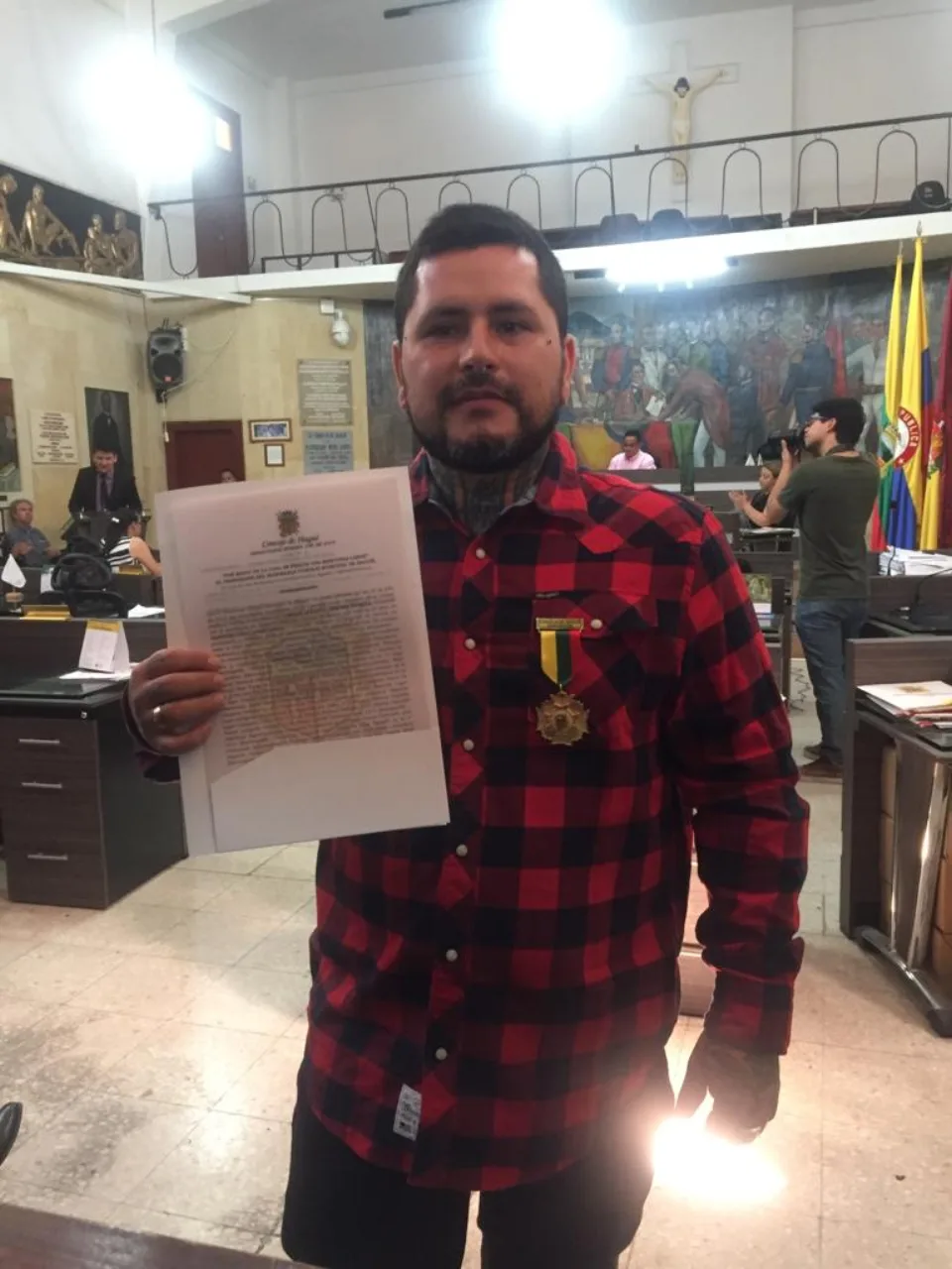 Dymon Jimenez in the council chamber holding the resolution, the medal pinned to his shirt