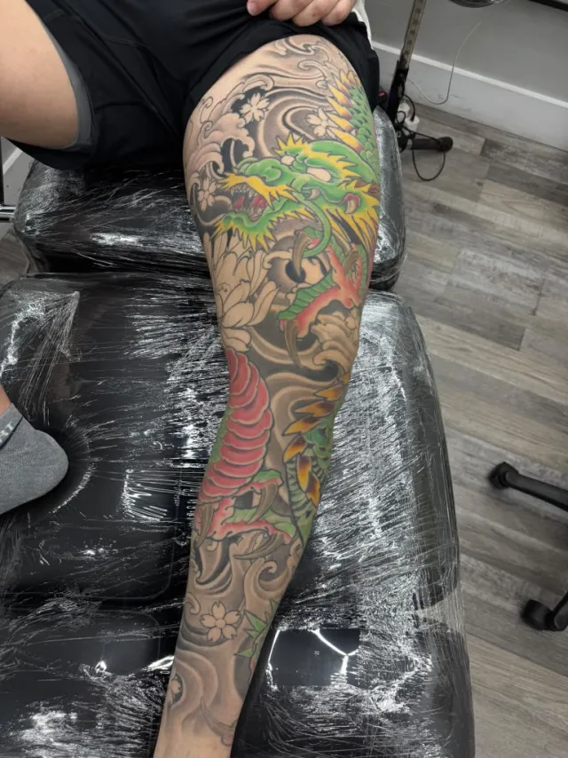 Client on the bench during a full-leg Japanese dragon session, protective film over the furniture